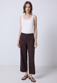 Pantalons cropped marron avec une coupe décontractée, fabriqués en tissu léger. Comprend des poches avant et une fermeture par bouton. Portés avec un débardeur blanc.