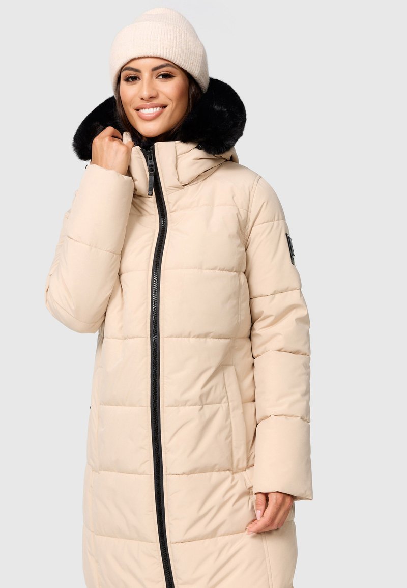 Beige gepolsterter Mantel mit schwarzem Reißverschluss und dickem Kunstpelzkragen, lange Ärmel und Seitentaschen. Das Model trägt eine leichte Strickmütze.