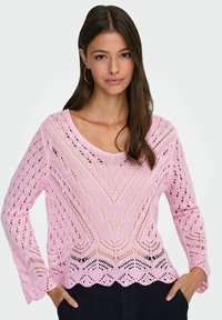 Femme portant un pull à manches longues en maille ouverte rose clair avec un ourlet festonné et un pantalon foncé, debout les mains dans les poches.