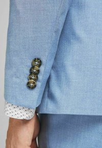 Ljusblå blazerärm med fyra dekorativa mörka knappar och en vit manschett med ett svart virvelmönster, som visar upp skräddarsydda design detaljer.