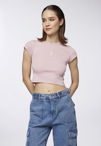 T-shirt cropped rosa chiaro con maniche corte, caratterizzato da un piccolo design di un orsetto ricamato sul petto. Indossato con pantaloni cargo in denim blu a vita alta.