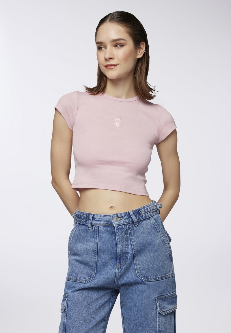 T-shirt cropped rosa chiaro con maniche corte, caratterizzato da un piccolo design di un orsetto ricamato sul petto. Indossato con pantaloni cargo in denim blu a vita alta.