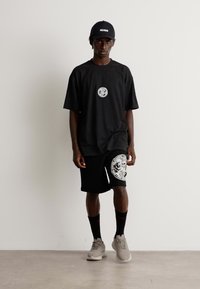 Mann i svart Hugo-cap, svart oversized t-skjorte og shorts med grafiske logoer, svarte sokker og lysegrå joggesko, står mot en hvit vegg.