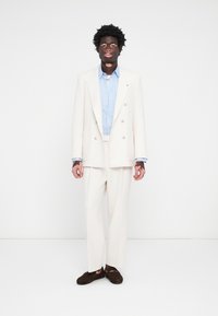 Cream double-breasted suit with a light blue shirt, relaxed fit pants, and dark brown shoes. Smooth fabric with subtle texture and detailing.