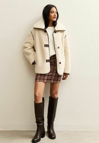 Chaqueta de shearling oversized en crema con acentos marrones, gran cuello, cierres de botones; suéter blanco de punto; falda de cuadros marrones; botas altas marrones hasta la rodilla.