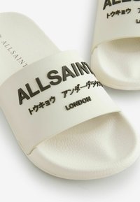 Bijele plažne sandale izrađene od teksturiranog gume s širokim remenom na kojem su crnim utisnutim slovima napisani: "ALLSAINTS", "LONDON" i japanski znakovi.