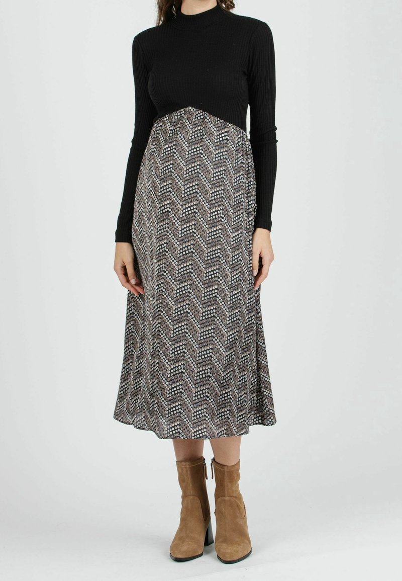 Haut à manches longues noir à rayures, associé à une robe au genou à motif en chevrons dans des tons sombres et avec des points multicolores. Bottes brunes à la cheville.