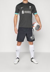 Harmaa Nike-jalkapallovarustus teal-vivahteilla, jossa Liverpool FC -logo, mustat shortsit ja valkoiset sukat. Pelaaja pitää käsissään kuvioitua jalkapalloa.