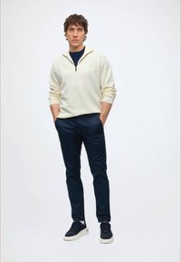 Ung mand stående med hænderne i lommerne iført en cremefarvet kvart-zip sweater, mørke bukser og marineblå sneakers mod en ensfarvet baggrund.