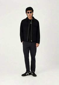 Casaco preto com zíper e dois bolsos frontais, usado sobre uma camisa preta. Calças escuras e ténis pretos com detalhes brancos. Óculos de sol redondos.