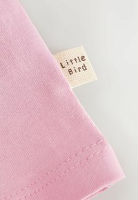 Coin de tissu rose avec une étiquette beige tissée portant l'inscription « Little Bird » cousue dans la couture sur un fond clair.