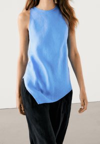 Light blue linen sleeveless top with a round neck and curved hem, paired with black loose-fitting pants. Smooth texture and simple design.
