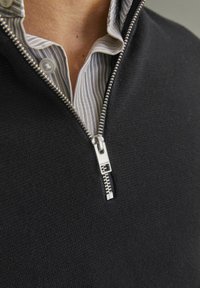 Melnā zipa džemperī ar sudraba rāvējslēdzēju, kas valkāts virs apakšsvārka ar zilu-striptētu kreklu. Uzsvars uz auduma tekstūru un kontrastējošiem rakstiem.