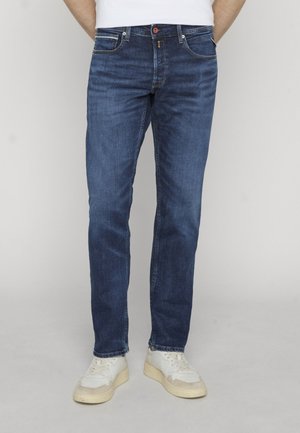 Man draagt slim-fit donkerblauwe spijkerbroek en witte sneakers, staand tegen een eenvoudige lichte achtergrond.