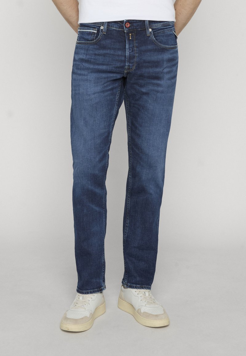 Homme portant un jean slim bleu foncé et des baskets blanches, debout devant un fond clair uni.