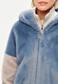 Giacca in pelliccia sintetica di colore azzurro chiaro con cappuccio. Presenta una chiusura frontale con zip, dettagli beige sulle maniche e una superficie morbida e testurizzata.