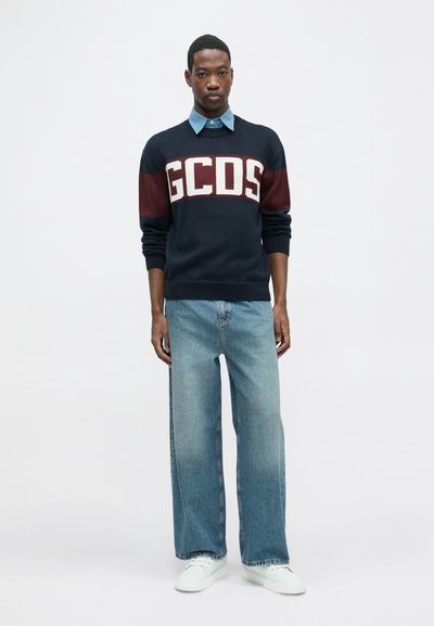 Tumesinine sviiter maroon triipudega ja valge "GCDS" logo, kombineeritud helesiniste denim-tekstuuriga teksade ja valgete tossudega.