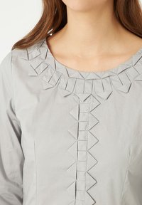 Blusa gris con un escote redondo, detalles en triángulo texturizados a lo largo del cuello y el frente, y un acabado de tela suave.