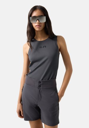 Vrouw met een mouwloos donkergrijs topje en donkergekleurde, hoog getailleerde korte broek met grote reflecterende zonnebril, staand tegen een effen achtergrond.