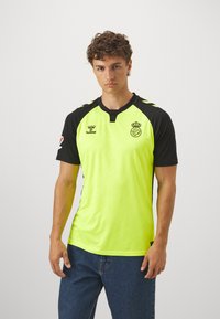 Maglia sportiva di un vivace giallo neon con maniche e colletto neri. Logo Hummel e stemma del club sul petto; motivo testurizzato sottile su tutta la superficie.