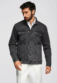 Giacca in denim nero con un taglio classico, dotata di due tasche sul petto, chiusura con bottoni e cuciture a vista. Indossata sopra una camicia bianca.