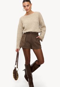 Pull beige duveteux, short en laine marron avec des plis, bottes marron au-dessus du genou et sac marron avec des accents dorés. Texture lisse partout.