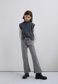 En ung person iført en grå ærmeløs turtleneck sweater over en hvid langærmet skjorte, grå flagrende jeans, sorte støvler og et beige bælte.