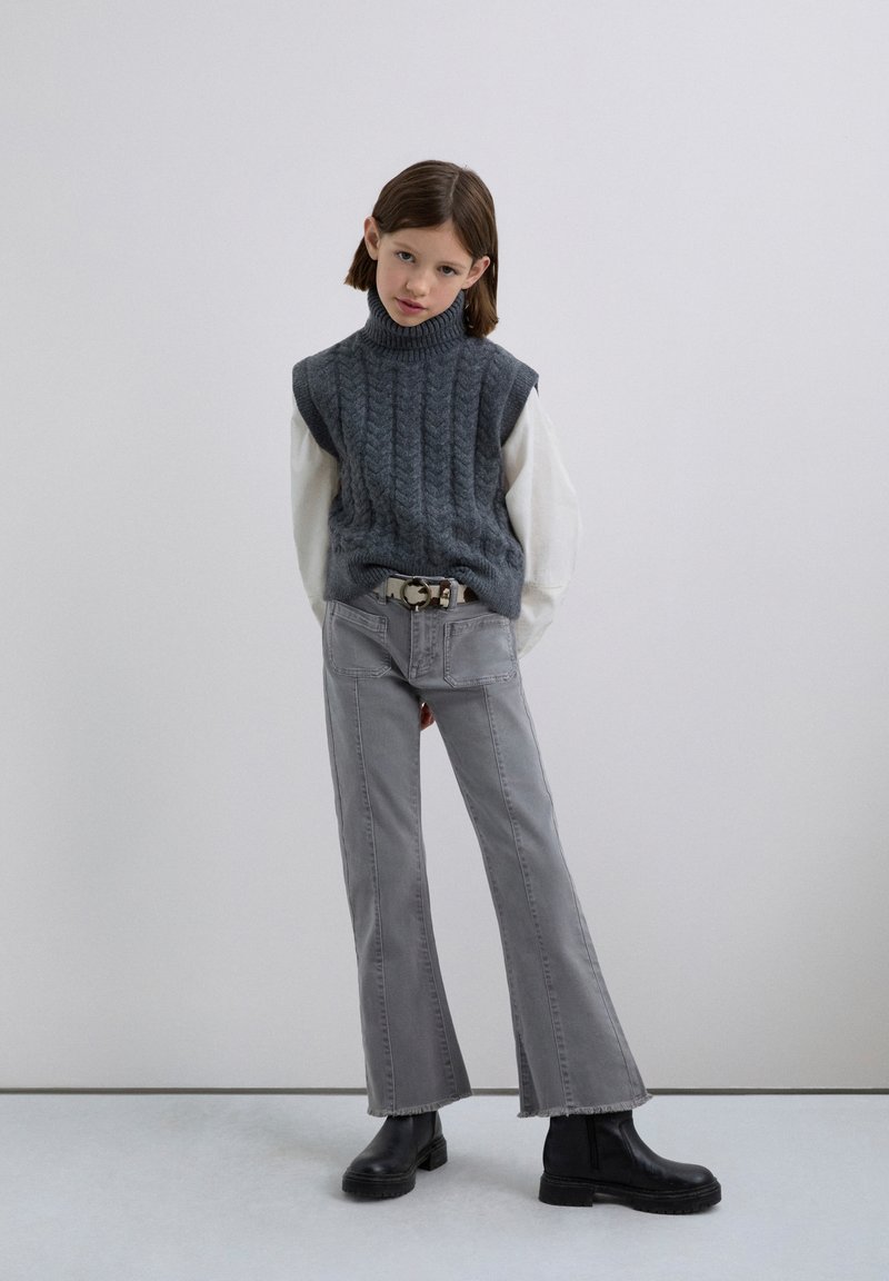 En ung person iført en grå ærmeløs turtleneck sweater over en hvid langærmet skjorte, grå flagrende jeans, sorte støvler og et beige bælte.