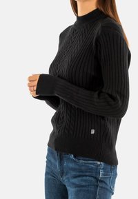 Pull en tricot noir avec texture côtelée, col montant et design en câble à motifs. Présente une petite étiquette de marque sur le côté inférieur.