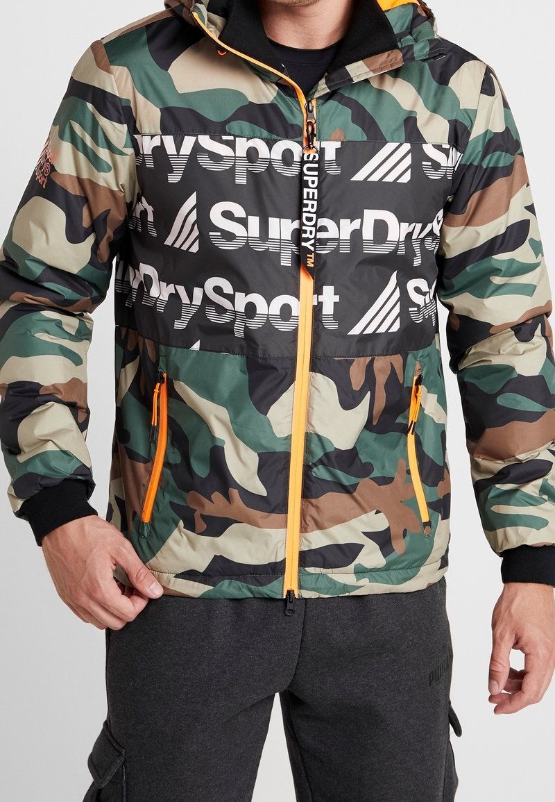 Uomo che indossa una giacca a stampa mimetica con pannello nero che presenta i loghi "SuperDry" e "DrySport", cerniera neon arancione e dettagli delle tasche, e pantaloni scuri.