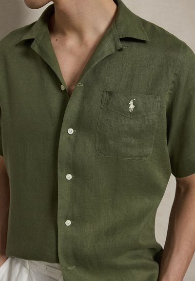 Homme portant une chemise kaki à manches courtes avec des boutons et une poche avant ornée d'un petit logo blanc de joueur de polo.