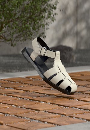 Μπεζ δερμάτινο σανδάλι με πόρπη, σχέδιο με λουράκια, μαύρη σόλα, εκτεθειμένο σε εξωτερικό χώρο πάνω σε επιφάνεια ξύλου με σχέδια και με φόντο θολό πράσινο τοπίο.