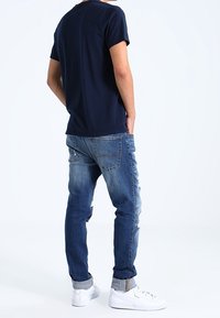 Man som står med ryggen vänd, iklädd en marinblå t-shirt, slitna blå jeans med uppvikta fållar och vita sneakers, mot en enfärgad vit bakgrund.