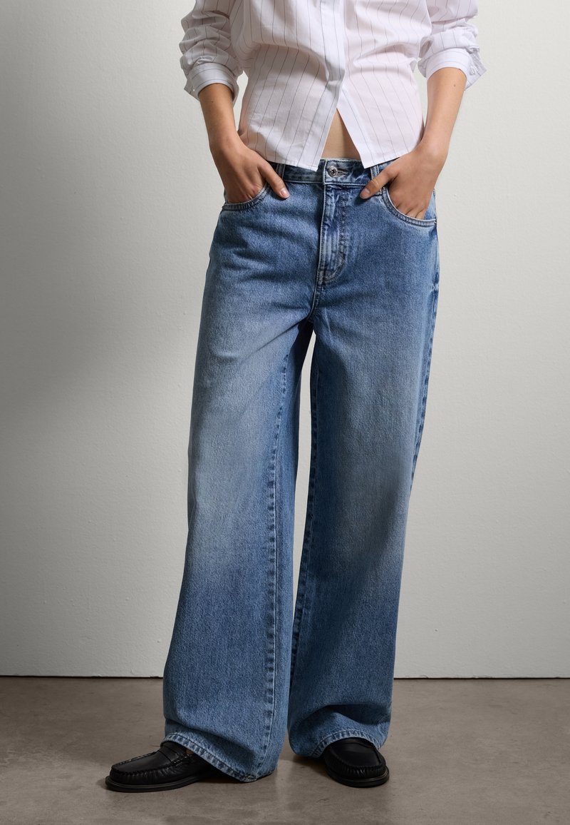 Jean large en denim bleu clair avec une finition délavée, dotés de poches avant et d'une fermeture à bouton. Associé à une chemise blanche à rayures et des mocassins noirs.