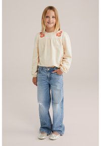 Top beige chiaro con texture e ricami floreali rossi, abbinato a jeans larghi di colore blu chiaro e scarpe slip-on argento scintillanti.