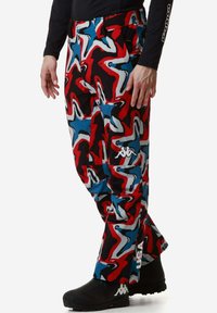 Pantaloni colorati a fantasia in rosso, blu e nero, con un design astratto fluido e un accento con logo sulla gamba sinistra. Il materiale sembra leggero.