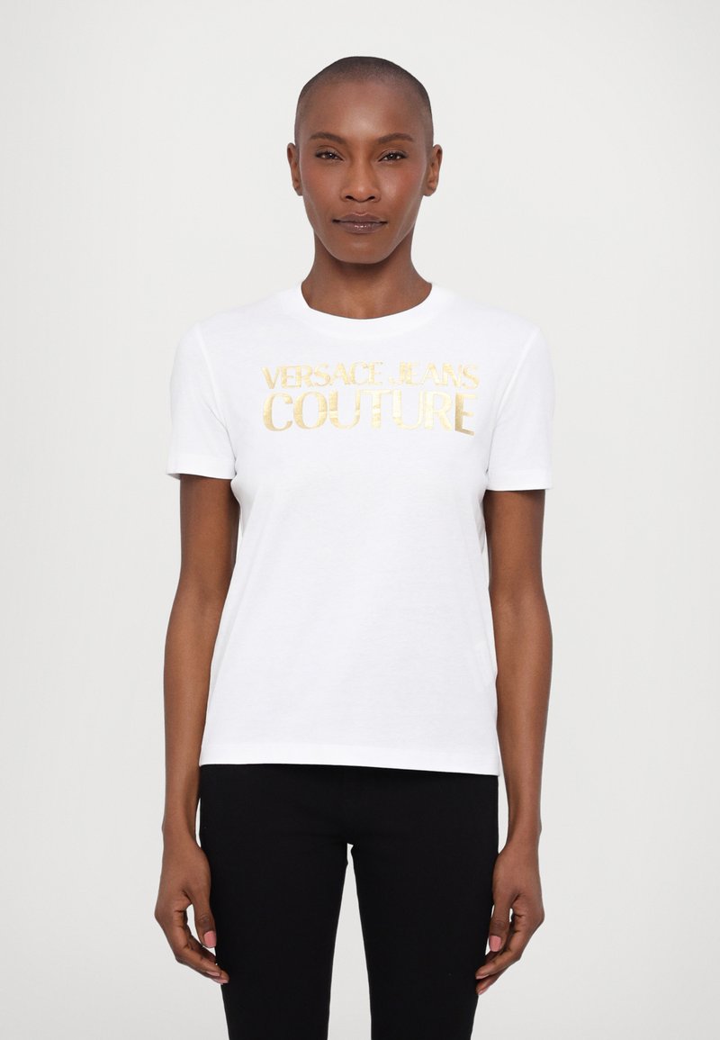 White cotton t-shirt featuring gold metallic text “VERSACE JEANS COUTURE,” round neckline, short sleeves, and a fitted silhouette.