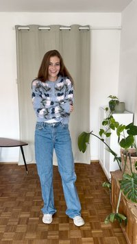 Suéter cinza tricotado com padrão floral, jeans de alta cintura azul claro e tênis brancos. Ambiente interno com plantas e chão de madeira.