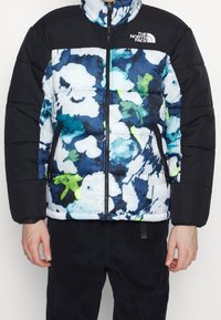 Chaqueta acolchada con una parte superior negra y un patrón floral en azul, blanco y verde en la parte inferior; incluye bolsillos con cremallera.