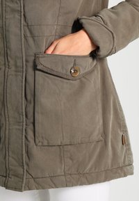 Main insérée dans une poche à rabat avec bouton sur une veste marron aux manches retroussées, montrant les détails de couture à l'avant et sur le côté.