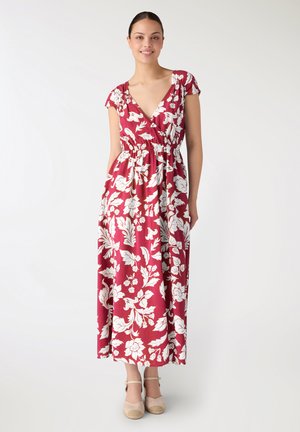 Donna in piedi che indossa un abito lungo rosso con motivo floreale bianco, maniche corte, scollo a V e scarpe beige con zeppa e cinturino alla caviglia, su sfondo semplice.