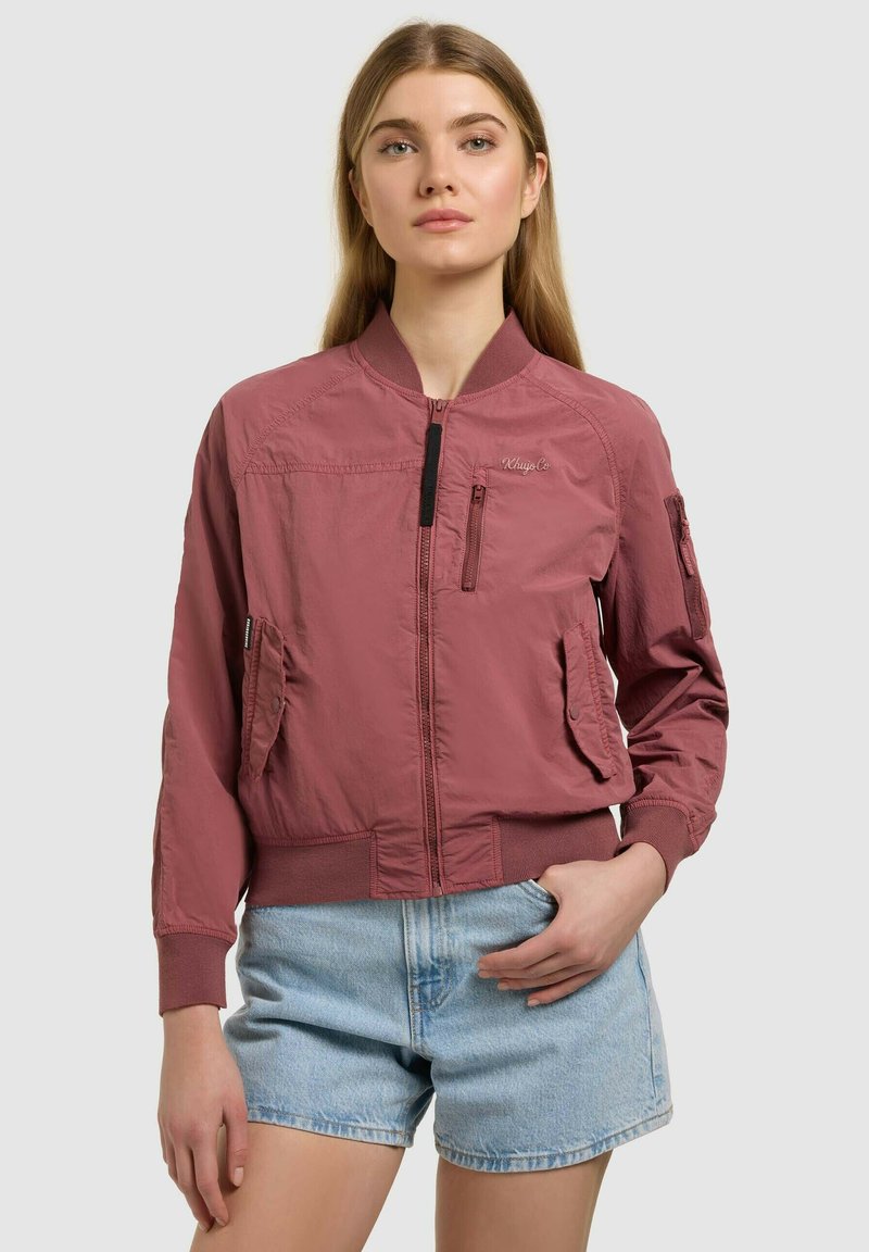Jeune femme portant une veste bomber mauve et un short en jean bleu clair, debout avec une main dans la poche sur un fond uni.