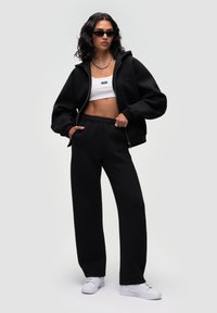 Sort zip-up hættebluse med logo, hvid crop top, sorte sweatpants og hvide sneakers. Inkluderer solbriller og en sølv halskæde.