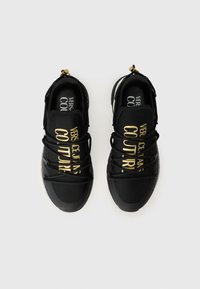 Des chaussures de sport noires au design épuré, dotées de lacets en élastique, portant le texte doré "VERSACE JEANS COUTURE", et une semelle en caoutchouc texturée.