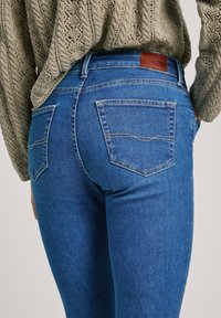 Vaqueros de denim azul oscuro con corte ajustado, que cuentan con dos bolsillos traseros y una etiqueta de cuero marrón encima de la cintura. Suéter de punto en color oliva arriba.