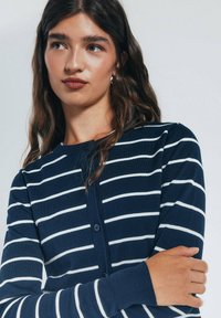 Námořnická modrá knoflíková cardigan s bílými vodorovnými pruhy, žebrovanými manžetami a měkkou texturou. Má kulatý výstřih a přiléhavý střih.