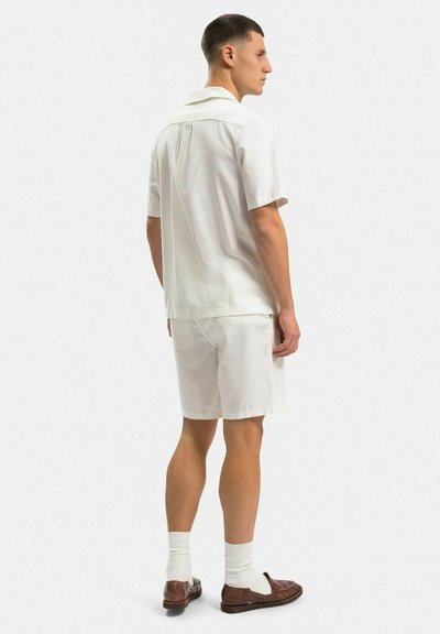 Hvid kortærmet skjorte og matchende shorts lavet af struktureret stof. Skjorten har en flad krave, rygfolder; bæres med brune slip-on sko.