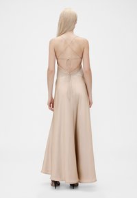 Robe longue fluide couleur nude avec un corsage ajusté, des bretelles croisées réglables et un dos ouvert, présentant une texture lisse et satinée.