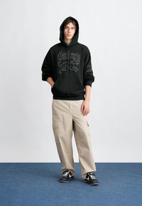 Sweatshirt preto com design gráfico, calças cargo bege e tênis pretos. O modelo está de pé sobre um tapete azul, com as mãos nos bolsos, olhando para a frente.