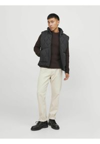Gepolsterte schwarze Weste mit hohem Kragen, kombiniert mit einem strukturierten dunkelbraunen Pullover, beigen Hosen und schwarzen Schuhen; neutraler Hintergrund.
